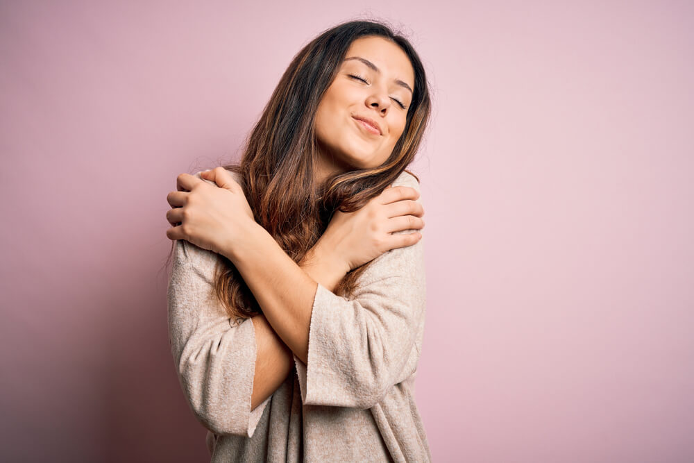 Young Beautiful Brunette Woman Wearing Casual Sweater Standing Over Pink Background Hugging Oneself