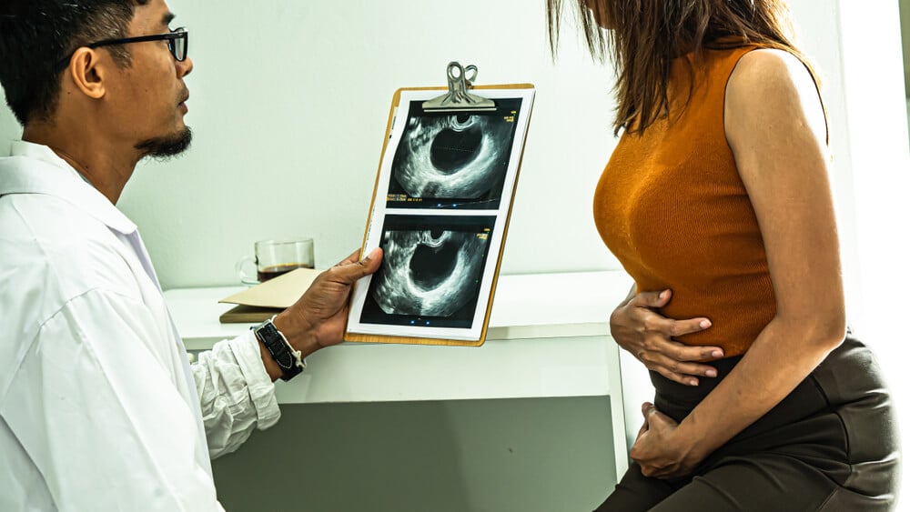 Doctor Holding X-Ray Film and Explaining About Uterus, Ovaries, Ovarian Cysts and Abnormalities in Cells, With a Female Patient Sitting in the Stomach Pain in Front